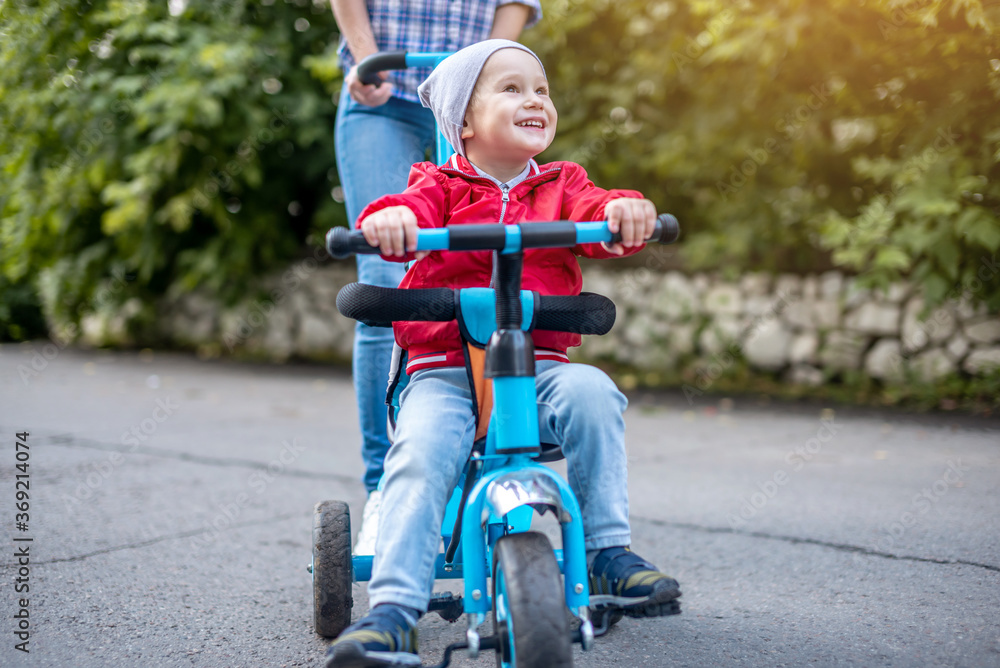 Young mother is pushing a child's tricycle with a toddler boy on a walk. Concept of learning to ride a bike and having fun with your family Young mother is pushing a child's tricycle with a toddler boy on a walk. Concept of learning to ride a bike and having fun with your family