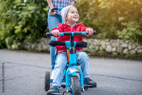Young mother is pushing a child's tricycle with a toddler boy on a walk. Concept of learning to ride a bike and having fun with your family