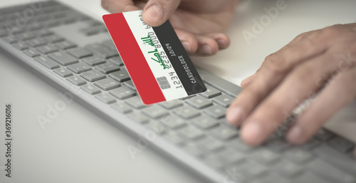 Man pays online with plastic bank card with printed flag of Iraq. Fictional numbers