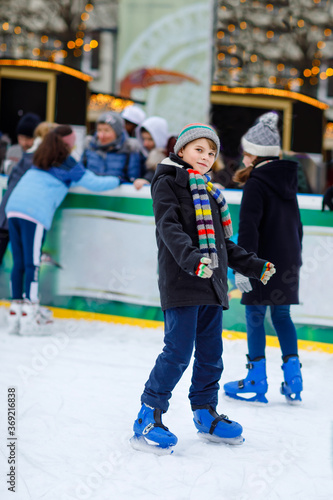 Two happy little kids girl and boy in colorful warm clothes skating on a rink of Christmas market or fair. Healthy children having fun on ice skate. Lot of people having active winter leisure.