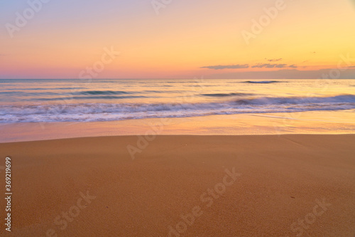 Colorful sunset at the tropical beach, sun behind clouds reflects on water and waves with foam hitting sand.