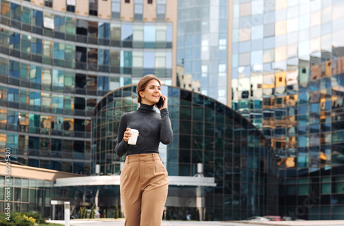 Young smiling business woman, with coffee in her hands, walks in the business part of the city and talks on a smartphone against the background of a business center