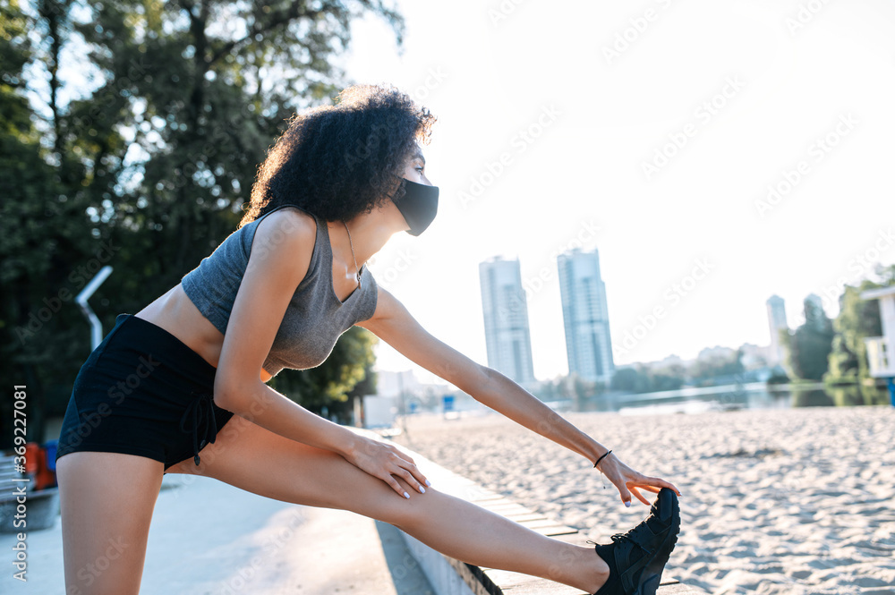 Sports training outdoor in safety during pandemic. An African-American woman with a medical mask on the face is stretching legs before outdoor workout. Side view with an urban landscape on background