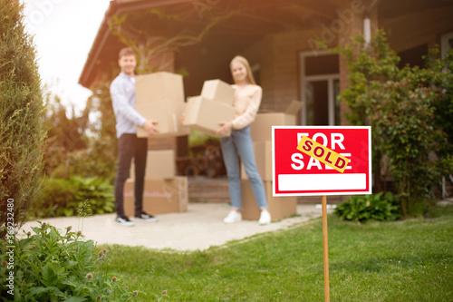 Young couple with boxes in front of their new house on moving day, focus on FOR SALE sign. Blank space