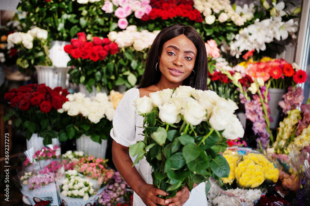 Beautiful african american girl in tender white dress with bouquet flowers in hands standing against floral background in flower shop.Black female florist.