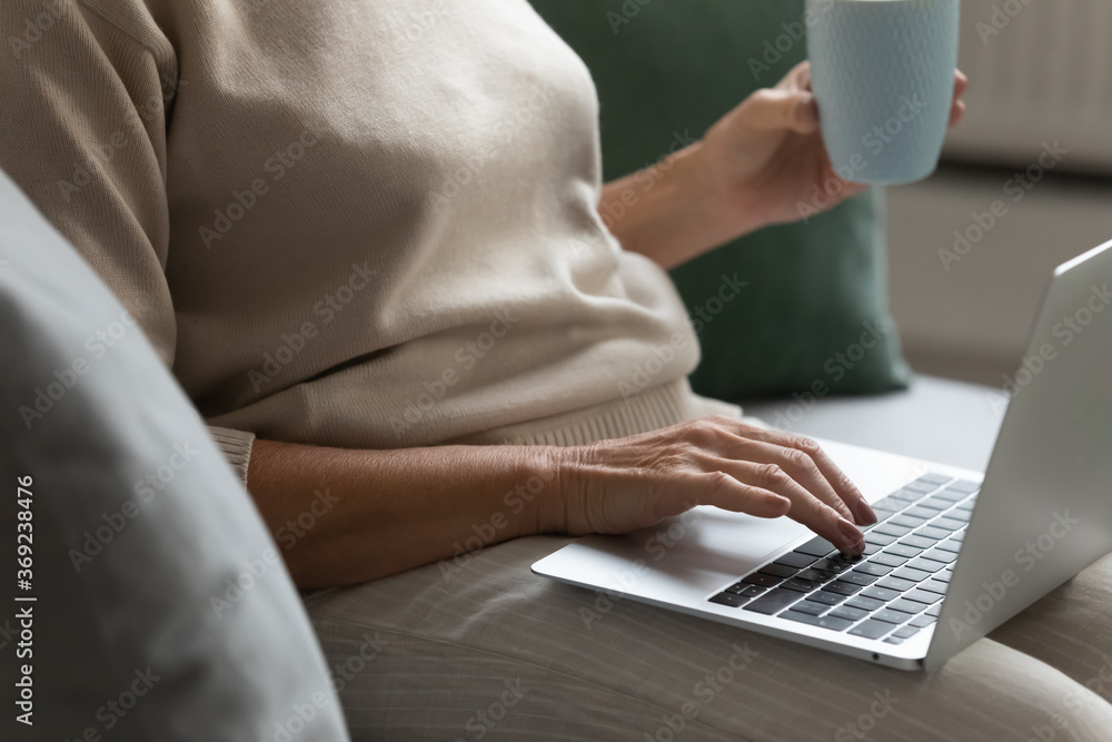 Naklejka premium Closeup view old woman sit on sofa hold cup put modern wireless computer on laps surfing cyberspace, read media news, communicates with grown up children on internet, buying in e-commerce site concept