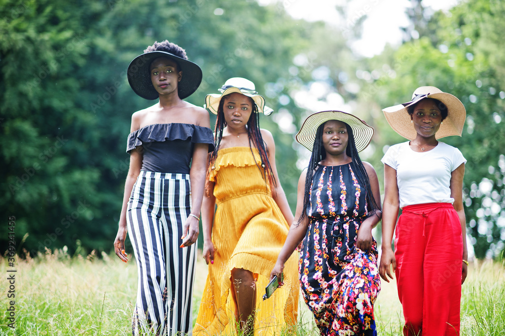 Group of four gorgeous african american womans wear summer hat spending time at green grass in park.
