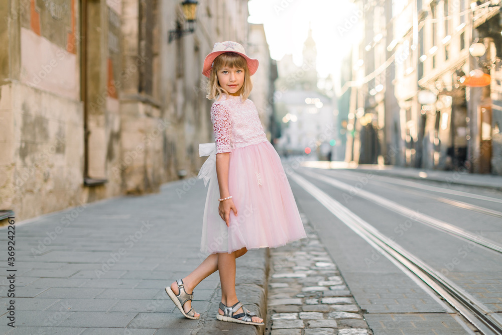 Stylish little girl wearing pink dress and hat, posing outdoors in the old European city, in sunny summer morning. Kid fashion concept
