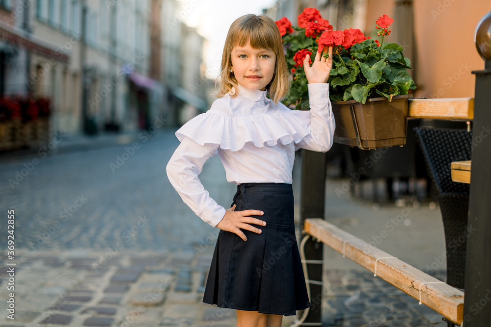 Closeup of little pretty blond Caucasian girl touching red flower pots decorations on summer day in old ancient European city