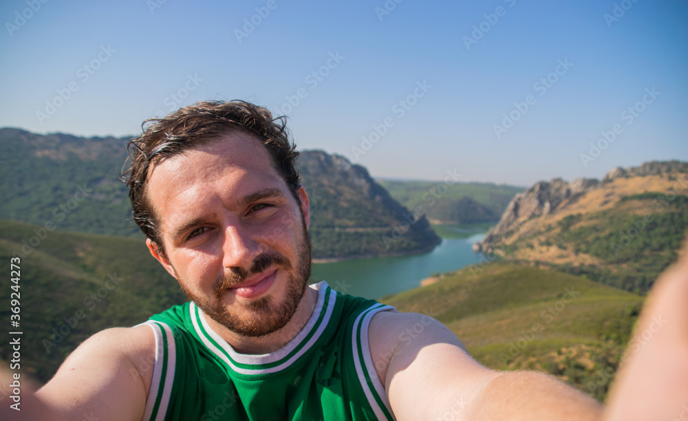 Photo of a young and attractive guy with beardtaking a selfie while travelling with a landscape behind him. Happy expression. Nature tourism