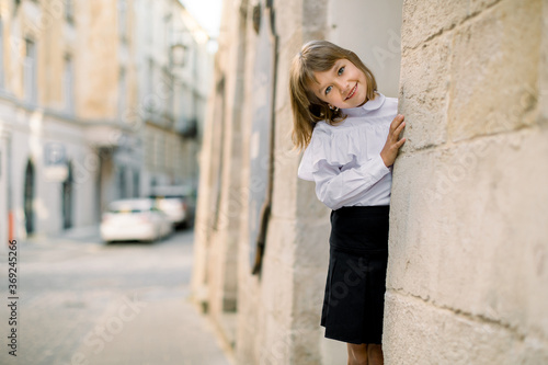 Cute little Caucasian blond girl in black and white clothes, posing to camera outdoors in the old city street, hiding her face behind the ancient building wall