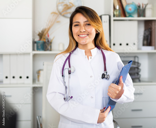 Young mexican female medic in uniform holding clipboard in doctor's office