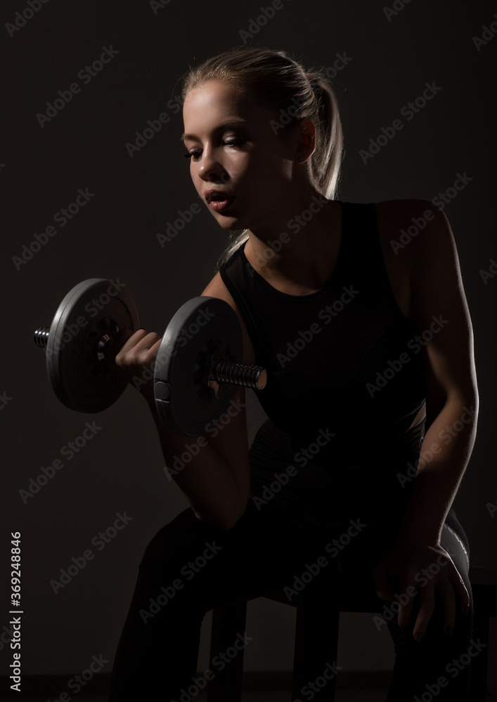 Obraz premium Fitness sexy girl with dumbbells on a dark background. Athlete doing exercises in the gym