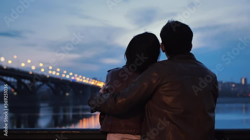 Rear view of a young couple hugging in the evening against a background of blured city lights. Lovers stand on the embankment and watch the evening city. The lights are reflected in the water. Slow