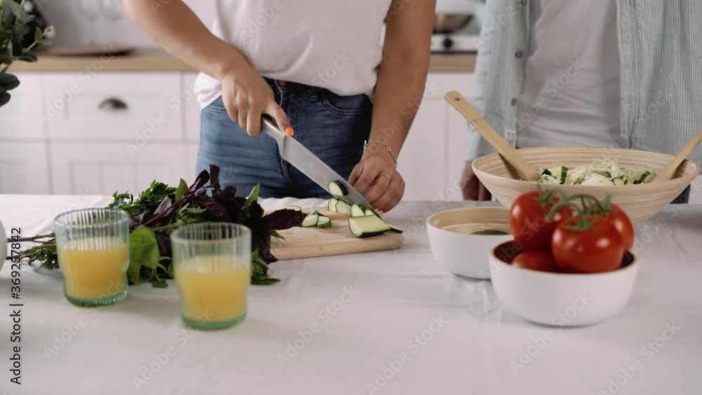 Cropped Close up of a Woman Chopping Cucumbers For a Vegetable Salad. Loving Husband is Helping a Wife to Cook a Healthy Meal. Family at Home. Cropped Close up of a Woman Chopping Cucumbers For a Vegetable Salad. Loving Husband is Helping a Wife to Cook a Healthy Meal. Family at Home.
