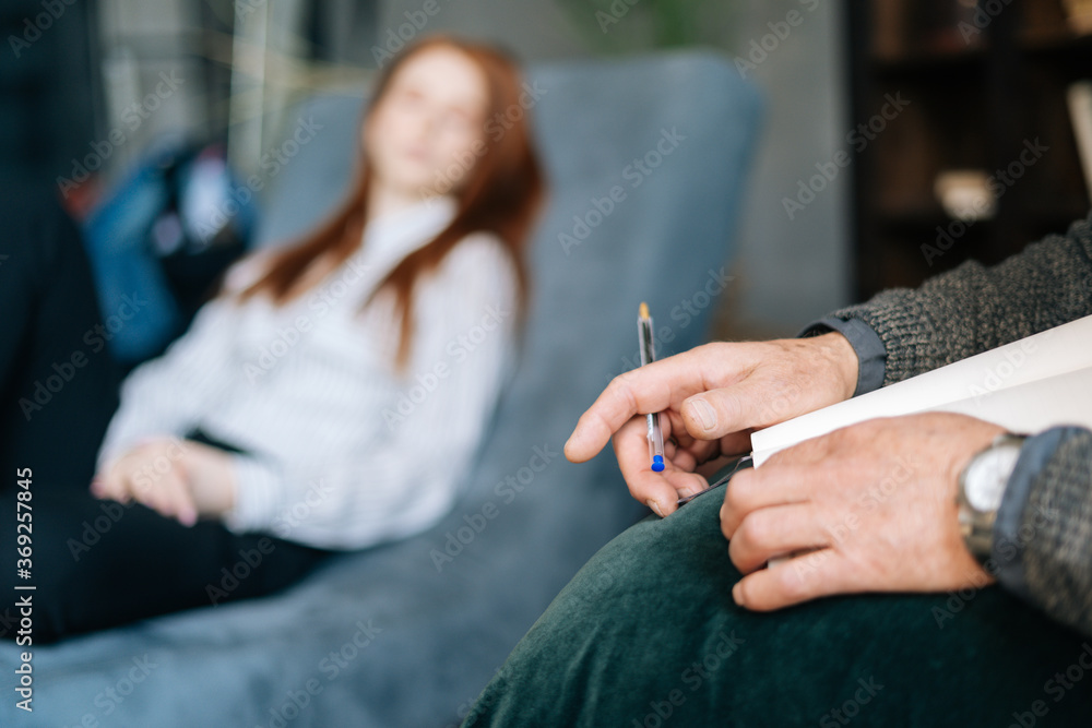 Close-up view of hands of mature man psychologist writing on clipboard while listening to depressed red-haired young woman patient during counseling therapy. Concept of psychological treatment.