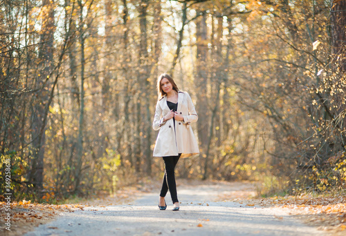 Smiling beautiful girl in a raincoat stands on the road in the autumn forest at sunset. Warm light. Yellow foliage © Александр Довянский