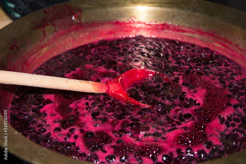 A wooden spoon is used to stir black currant jam boiling in a copper basin