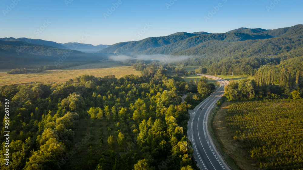 Idyllic landscape shot from above in early morning sunlight, meadows and forests with blue sky and a road leading to a mountains on the horizon
