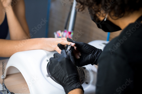 manicurist master in black gloves and mask is making manicure with a manicure drill apparatus in the nail salon