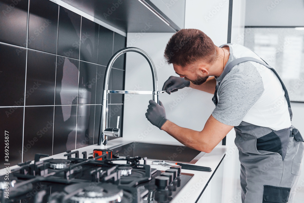 Young professional plumber in grey uniform fixing water tap on the kitchen Young professional plumber in grey uniform fixing water tap on the kitchen