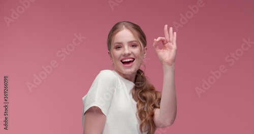 Attractive red-haired Girl is showing Okey to the Camera, smiling charmingly. Sunshiny young Woman showing great Emotions on pink Background on Studio. Cool Girl. Positive Lifestyle. Happy Woman. App. photo