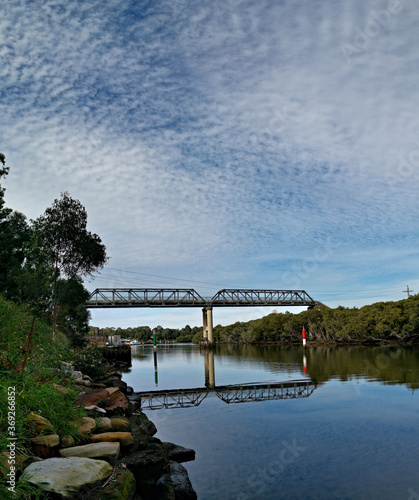 Beautiful view of a river with reflections of a tall pedestrian and water pipe bridge, trees, deep blue sky and puffy clouds on water, Parramatta river, Rydalmere, Sydney, New South Wales, Australia 