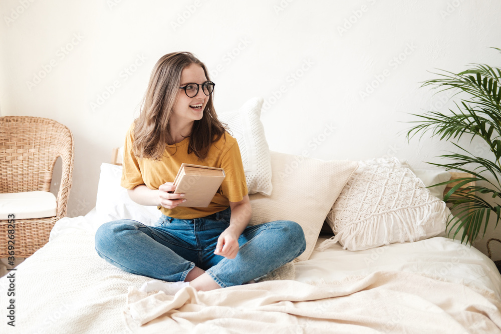 Young beautiful girl student in glasses, an orange T-shirt and blue jeans sits on bed with pillows and reads paper book. Self-education concept at home during quarantine. Home schooling, hobby. Young beautiful girl student in glasses, an orange T-shirt and blue jeans sits on bed with pillows and reads paper book. Self-education concept at home during quarantine. Home schooling, hobby.
