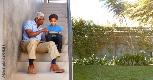 Grandfather With Grandson Sitting On Steps Outdoors At Home Using Digital Tablet