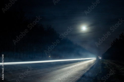 Light trails from vehicles traveling on rural road, surrounded by two rows of trees, late at night with moon on sky.
