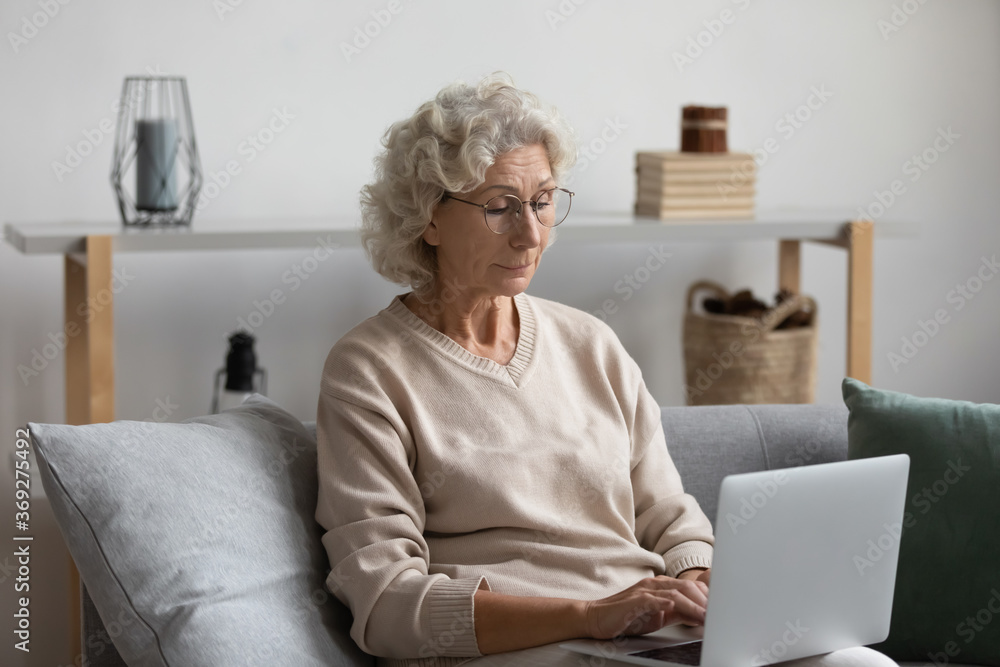 Serious old woman put computer on lap typing on device sit on sofa in living room. Retired female do telecommuting work, part time job from home on retirement, older generation and modern tech concept Serious old woman put computer on lap typing on device sit on sofa in living room. Retired female do telecommuting work, part time job from home on retirement, older generation and modern tech concept