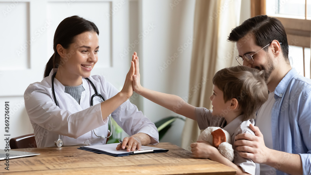 Obraz premium Happy young pediatrician in white uniform giving high five to small child boy sitting on fathers lap, celebrating good treatments results or encouraging, friendly doctor patient relations concept.