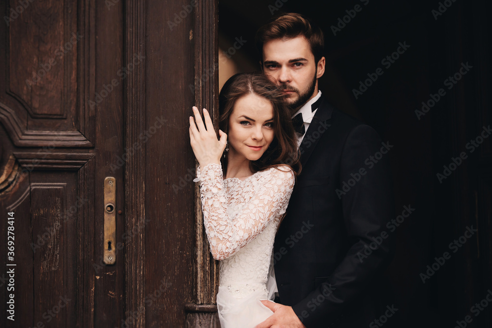 Beautiful newlyweds hugging near the ancient door. Wedding portrait of a stylish groom and a young bride near old house in in a European town