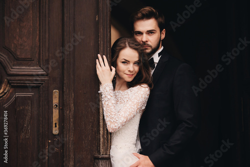 Beautiful newlyweds hugging near the ancient door. Wedding portrait of a stylish groom and a young bride near old house in in a European town