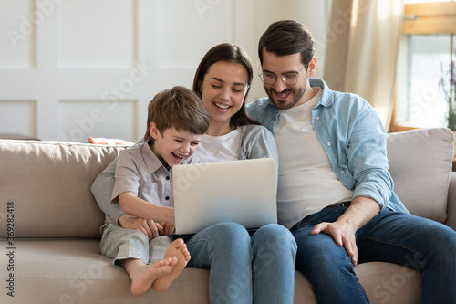 Happy young woman holding computer on laps, watching funny comedian movie video or shopping online with loving husband and adorable little child son, relaxing together on couch in living room.