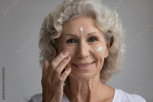 Close up face elderly happy attractive woman, 65s grey haired female smiling looking at camera feels satisfied applies moisturizer facial cream serum for senior skin, age defense treatment ad concept photo
