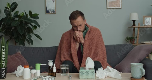 Bearded guy covered in blanket being ill while sitting on sofa at home. Man in 30s coughing loudly and s while getting sick and taking cure at home. Concept of healthcare and illness photo