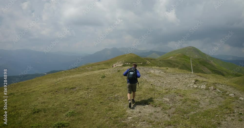 Aerial shot of a carefree man tourist with backpack is hiking on the path in the middle of hills surrounded by green nature in a magical and relaxing place.