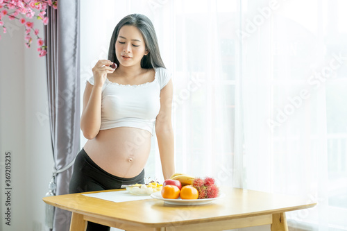 Soft blur of beautiful pregnant woman hold berry and stand near table with various types of fruits in front of white curtain with morning light. Concept of good and healthy food for pregnant people.