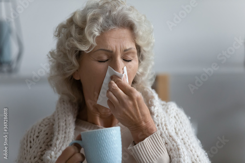 Close up unhealthy elderly senior woman caught a cold covered in warm knitted plaid use tissue wiping sneezing blowing runny nose. Sick granny treated at home holds cup takes drinks antiviral beverage
