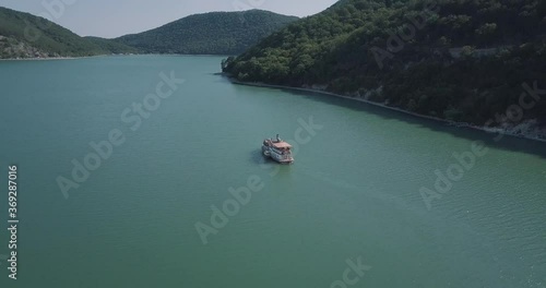 aerial video shooting from a drone flying behind a retro ship with passengers floating on a lake against the background of mountains and forests