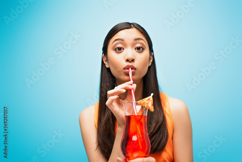 attractive asian girl holding glass and drinking cocktail through straw on blue