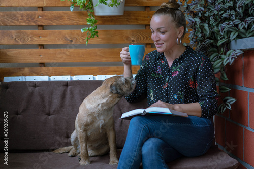 Young pretty woman sitting at balcony  drinking coffee and reading a book, enjoying with her dog border terrier