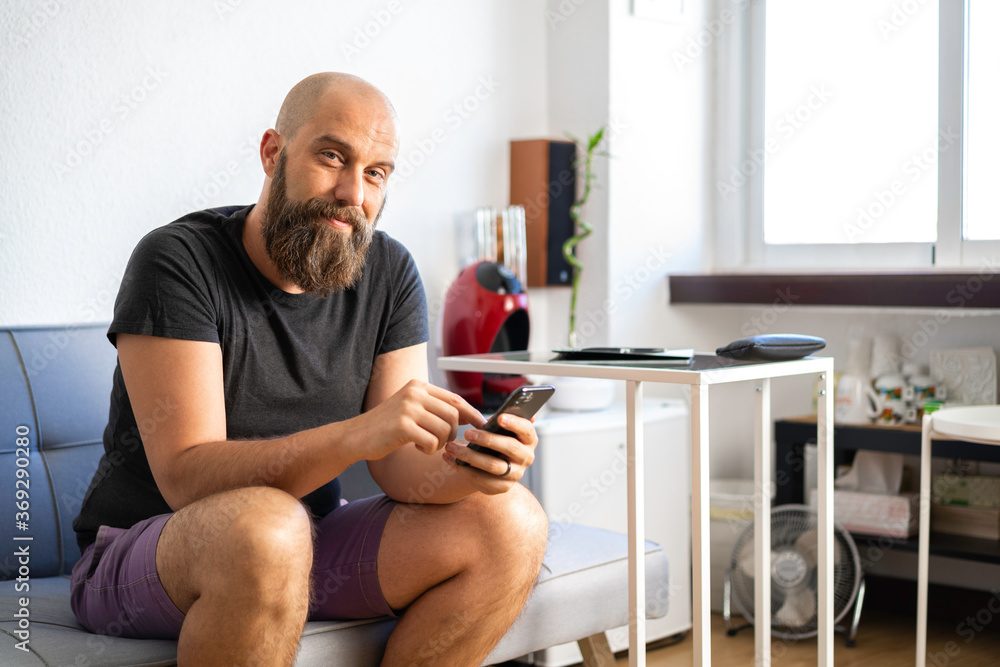 Hombre calvo con camiseta gris oscura, pantalón corto púrpura, y barba, mirando a cámara, sonriendo, con smartphone, sentado en un sillón en su oficina, con luz de día que entra por la ventana Hombre calvo con camiseta gris oscura, pantalón corto púrpura, y barba, mirando a cámara, sonriendo, con smartphone, sentado en un sillón en su oficina, con luz de día que entra por la ventana