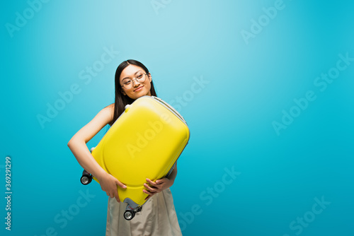 positive asian woman in glasses holding yellow luggage on blue