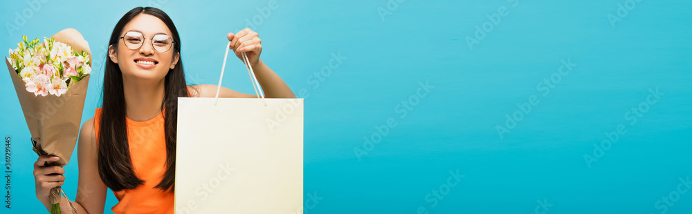horizontal crop of happy asian woman in glasses holding shopping bag and flowers on blue horizontal crop of happy asian woman in glasses holding shopping bag and flowers on blue