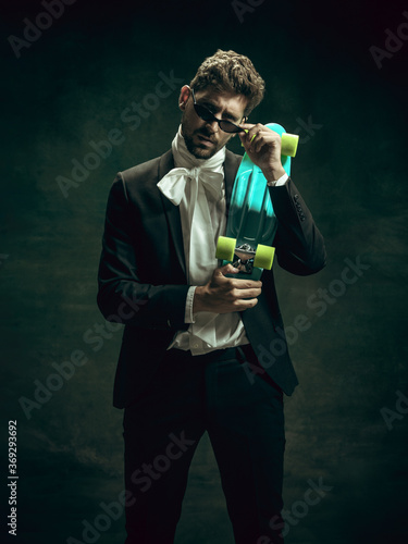 Skateboard urban style. Young man in suit as Dorian Gray isolated on dark green background. Retro style, comparison of eras concept. Beautiful male model like classic literature character, old