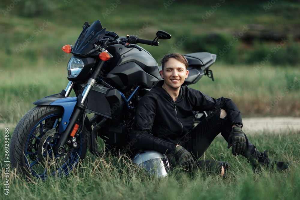 Image of biker sitting on the grass in front of motorcycle in sunset on the country road.