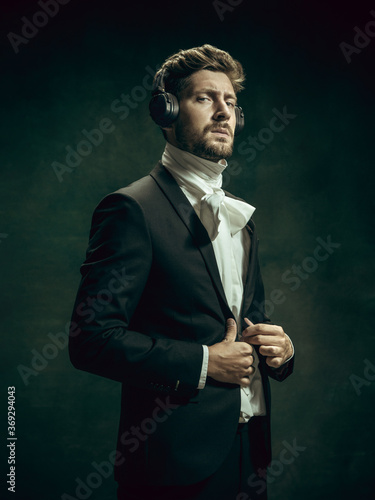 Music listening. Young man in suit as Dorian Gray isolated on dark green background. Retro style, comparison of eras concept. Beautiful male model like classic literature character, old-fashioned. photo