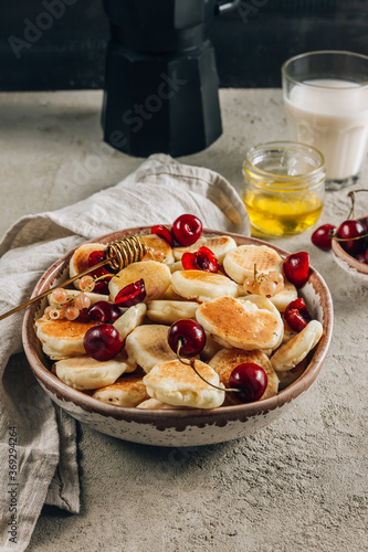 Trendy home breakfast with tiny pancakes in a bowl with honey and cherries on a concrete table. Selective focus. Vertical photo